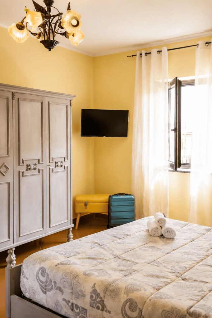 Armadio grigio a tre ante, televisore a parete e finestra con tende chiare accanto al letto matrimoniale
