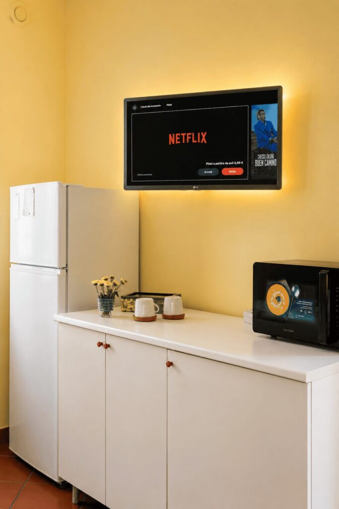 Credenza bianca con frigorifero, forno a microonde e tazze, sormontata da un televisore acceso