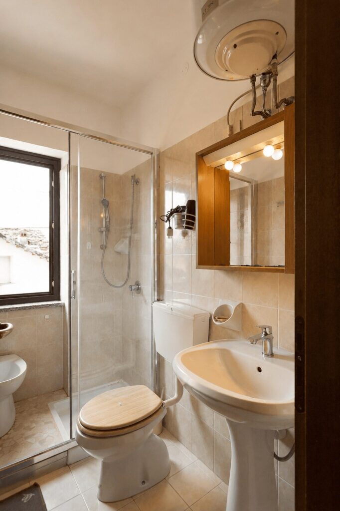 Bagno con box doccia in vetro, lavabo a colonna, specchio contenitore in legno e finestra sui tetti
