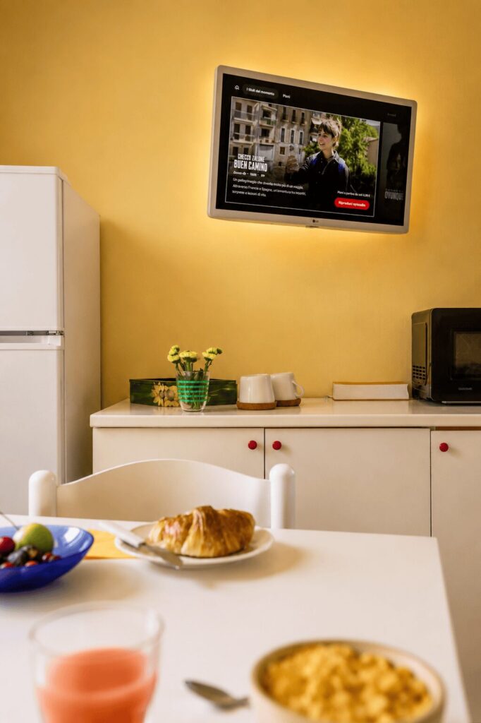 Cornetto, succo e cereali in primo piano sul tavolo, con il televisore acceso sopra la credenza