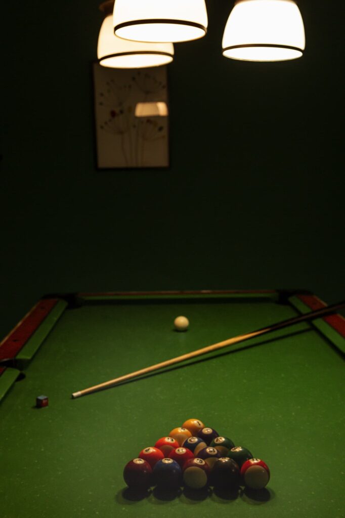 Sala giochi con tavolo da biliardo verde, triangolo di bilie e stecca, illuminata da lampade a sospensione