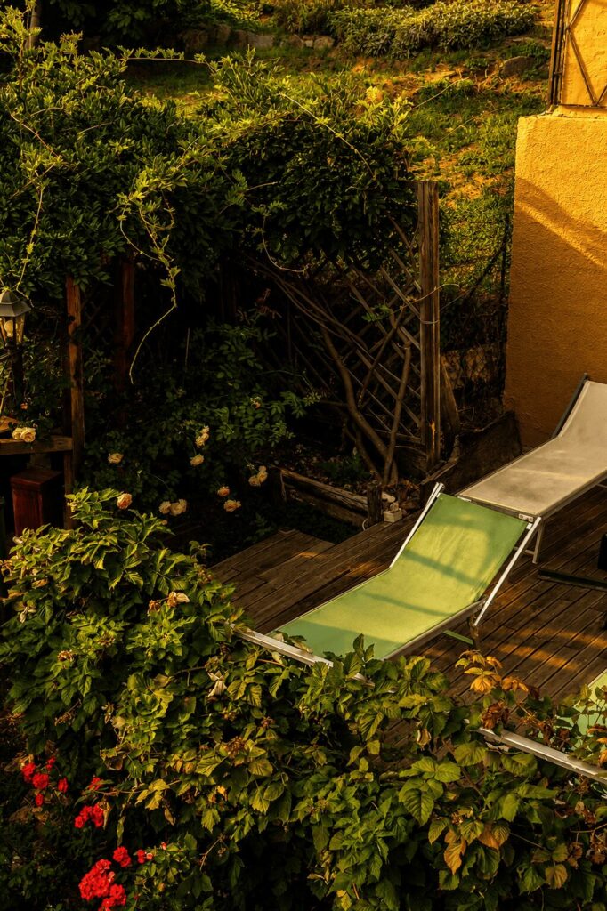 Due lettini prendisole su una pedana di legno, circondati da siepi, gerani rossi e un lampione da giardino
