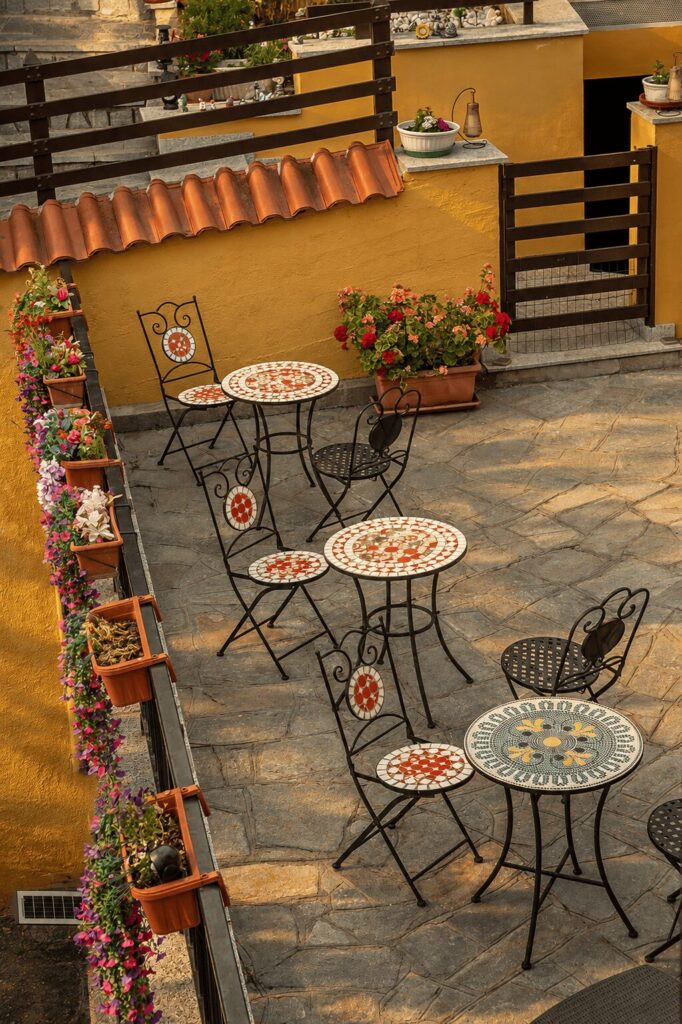 Terrazza in pietra con tavolini e sedie in ferro battuto decorati a mosaico, vasi di gerani e petunie lungo la ringhiera