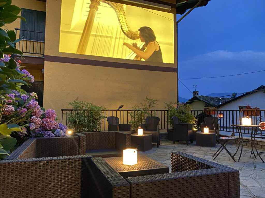 Terrazza al tramonto con divanetti in rattan, lanterne accese, ortensie in fiore e un maxi schermo che proietta un concerto di arpa