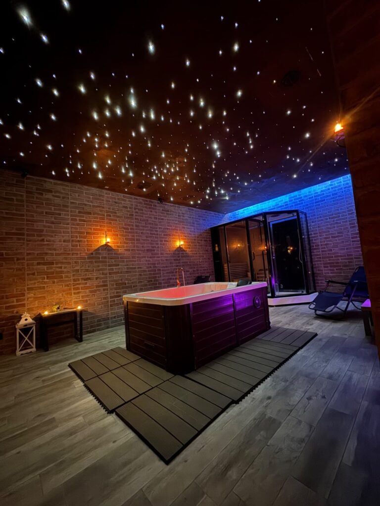 Vasca idromassaggio illuminata di viola su pedana in legno, muri in mattoni e soffitto a cielo stellato